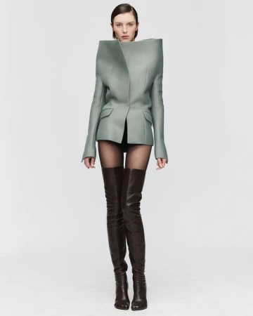 Fashion model in studio wearing an avant-garde blazer and thigh-high boots against a stark white background in a fashion editorial shot.