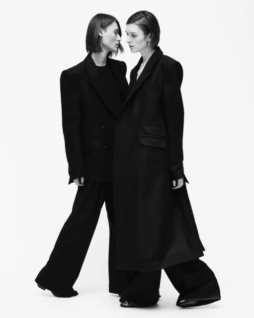 Monochrome fashion shot featuring two female models in tailored black suits against a bright backdrop, emphasizing shadows and modern style.