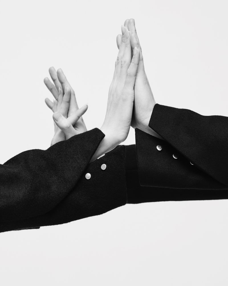 Monochrome studio shot showcasing two individuals in black jackets, their hands touching, set against a bright white backdrop. 