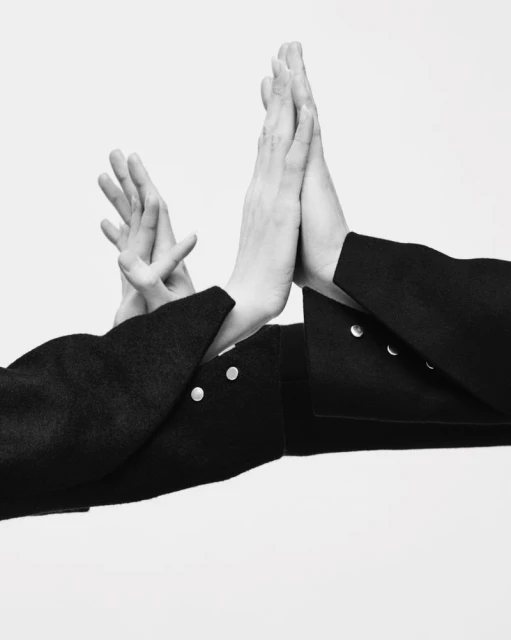Monochrome studio shot showcasing two individuals in black jackets, their hands touching, set against a bright white backdrop. 