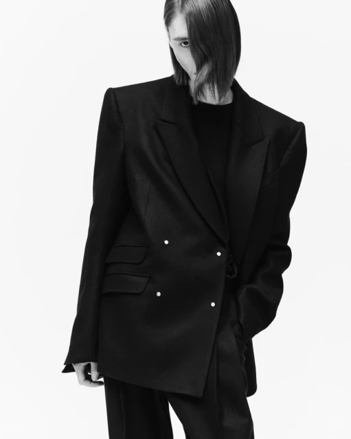Studio shot of a model wearing a black double-breasted blazer and black pants against a white backdrop with asymmetrical hair.