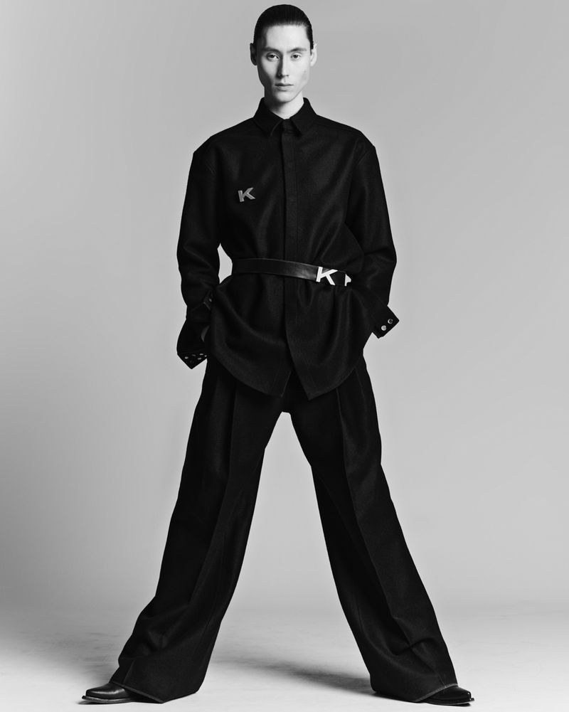 Monochrome studio shot of a model in an oversized black button-down shirt, wide-leg trousers, and belt with metal initial buckle.