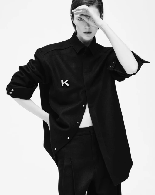 Black and white portrait of a slim adult fashion model in a black shirt and pants, striking a confident pose in a photo studio.