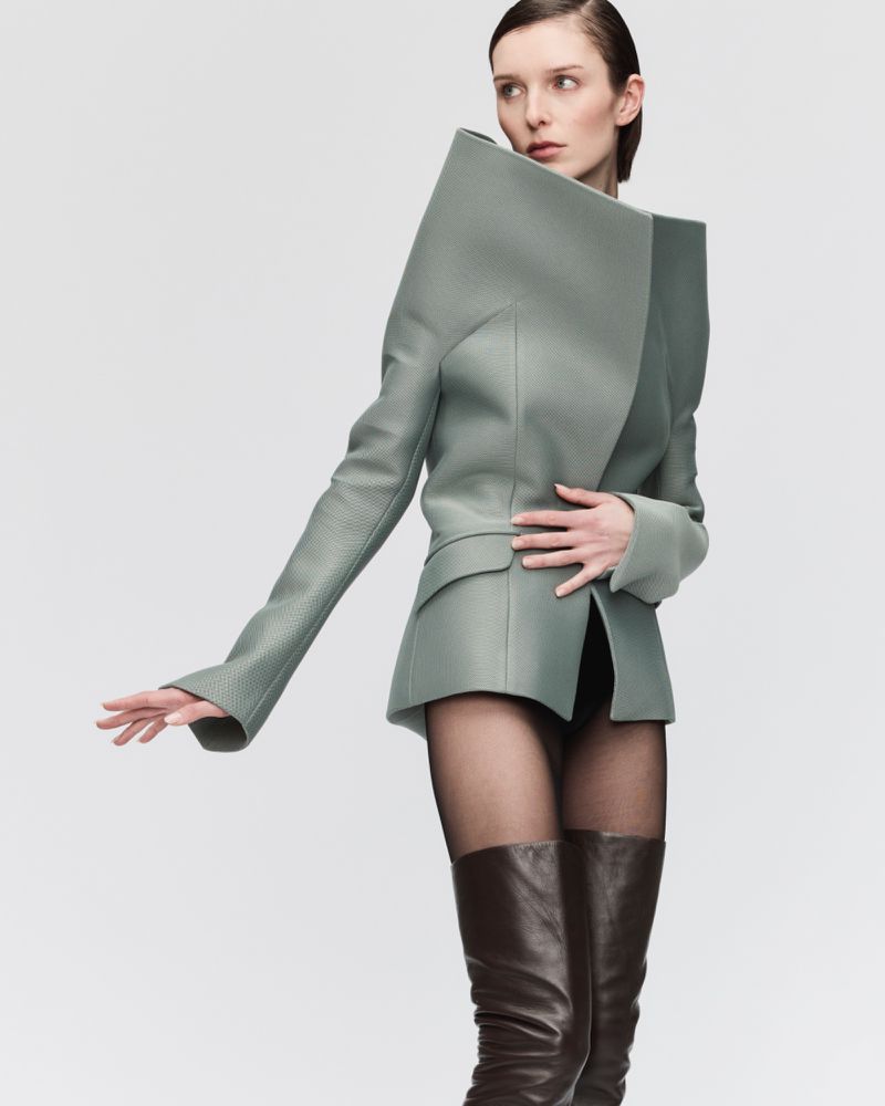 Fashion model in a light green blazer with exaggerated shoulders, black tights, and brown leather boots against a neutral backdrop.