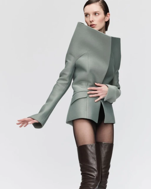 Fashion model in a light green blazer with exaggerated shoulders, black tights, and brown leather boots against a neutral backdrop.