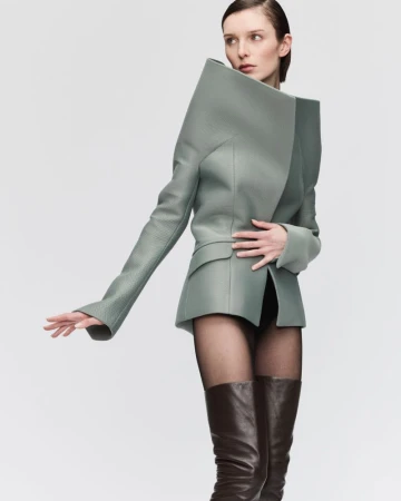 Fashion model in a light green blazer with exaggerated shoulders, black tights, and brown leather boots against a neutral backdrop.
