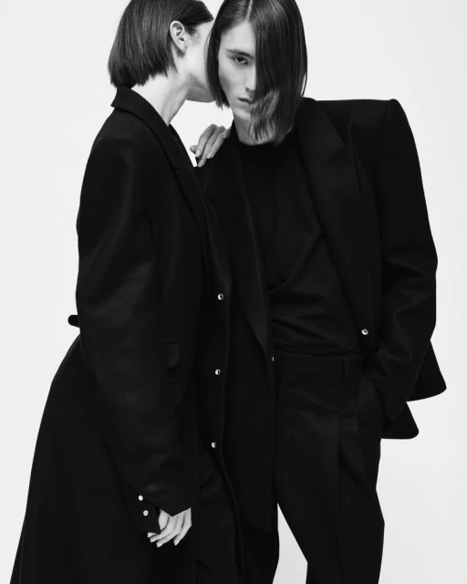 Black-and-white studio shot of an adult woman in a coat whispering to an adult male in a suit, both with serious expressions.