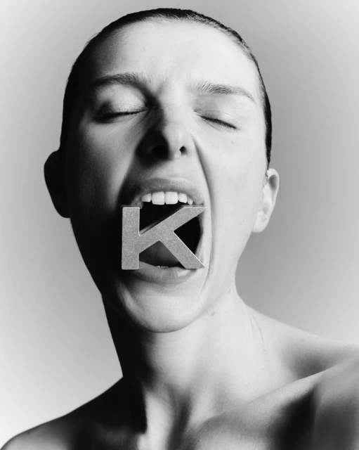 Black and white portrait of a Caucasian woman with her eyes closed and mouth open, holding a large letter K in her mouth.