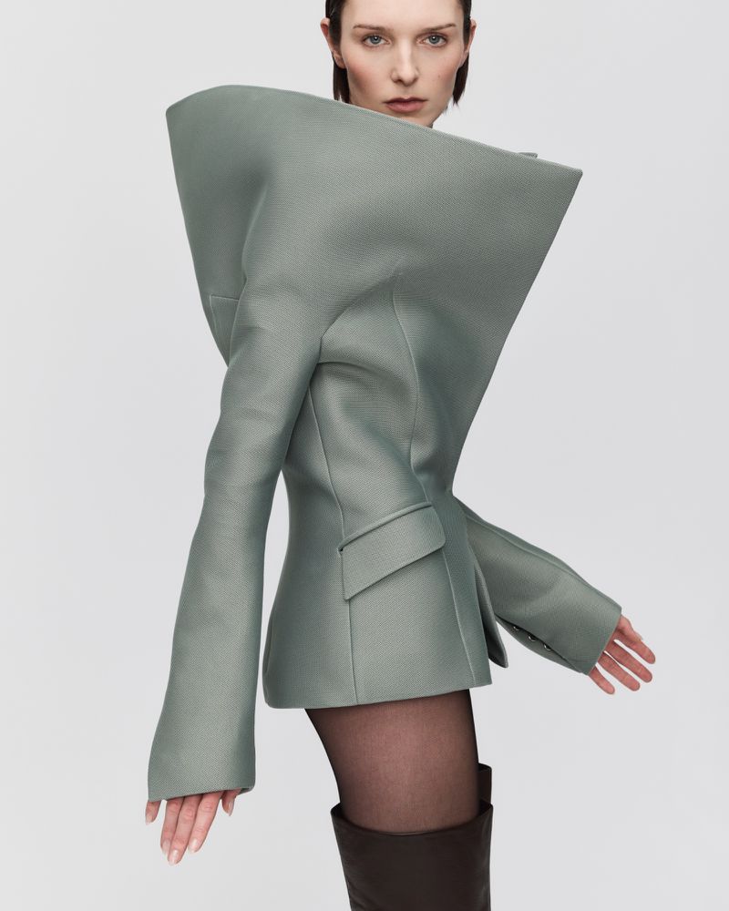 Model wearing a structured sage green high-collar jacket with exaggerated shoulders and tall brown boots against a light grey background.
