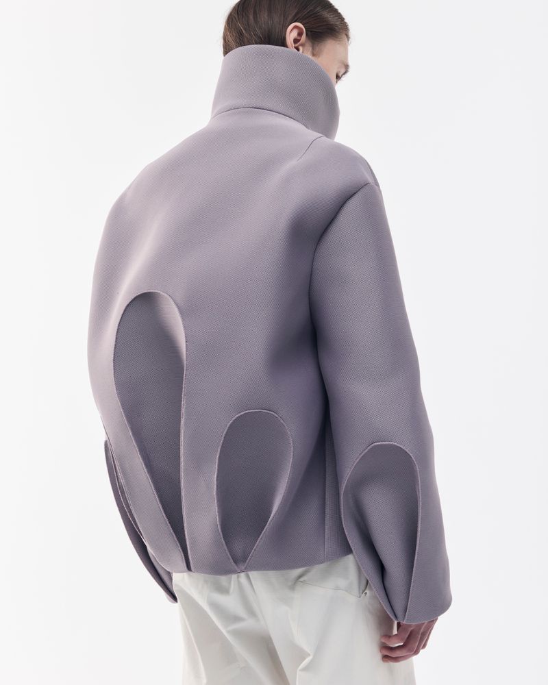 Person from the back wearing an oversized lavender jacket with large cut-out details and light-colored trousers against a white backdrop.
