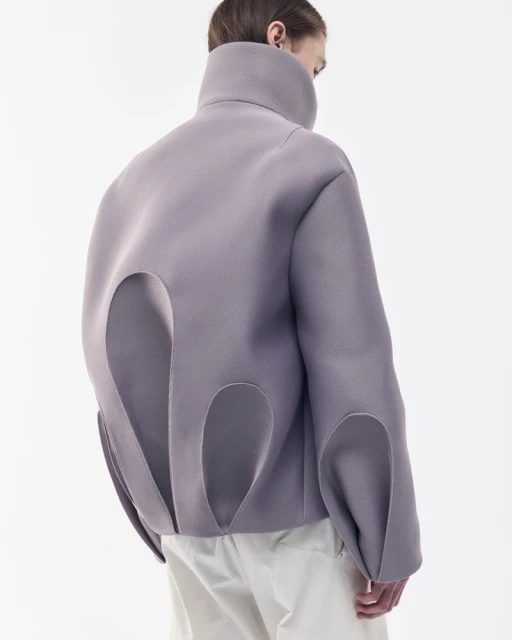 Person from the back wearing an oversized lavender jacket with large cut-out details and light-colored trousers against a white backdrop.