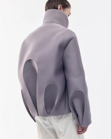 Person from the back wearing an oversized lavender jacket with large cut-out details and light-colored trousers against a white backdrop.