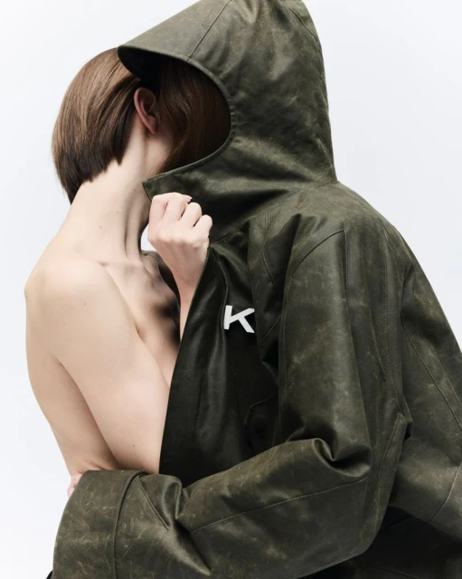 Fashion shot featuring an adult with short hair and a partial view of a green hooded jacket with the letter K on a white background.