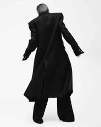 Model seen from behind wearing a long black coat with structured shoulders against a seamless white background.