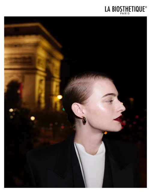 Portrait of a model with dark lipstick and earrings, wearing a black suit jacket, with the Arc de Triomphe in the background at night.