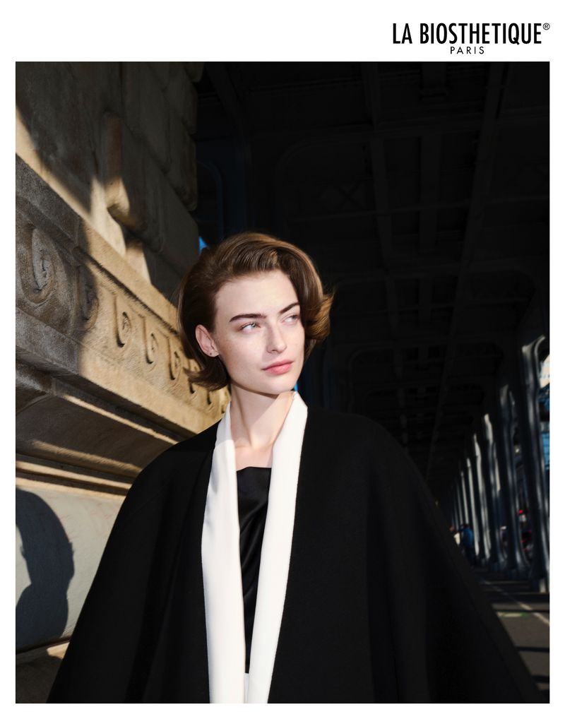 Elegant woman in black and white fashion poses against a dark urban architectural backdrop with diffused sunlight casting shadows.