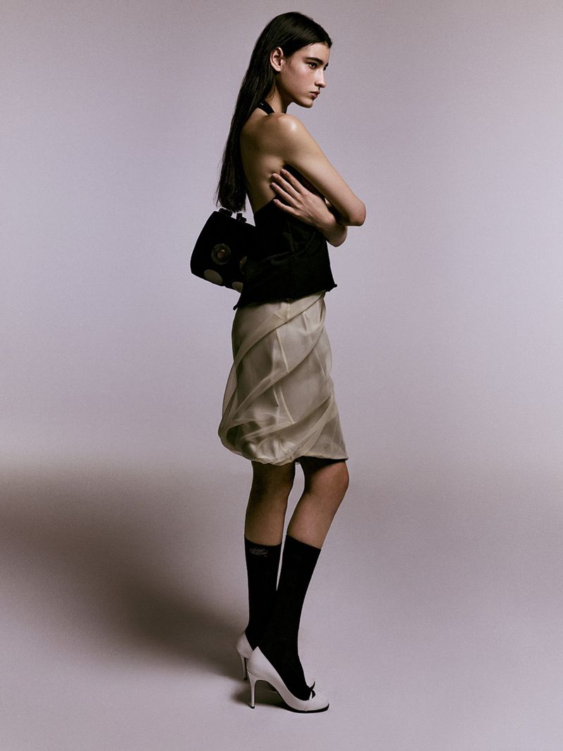 A slender woman poses in a fashion shot, showcasing a black halter top, beige skirt, black socks, and white pumps against a neutral backdrop.