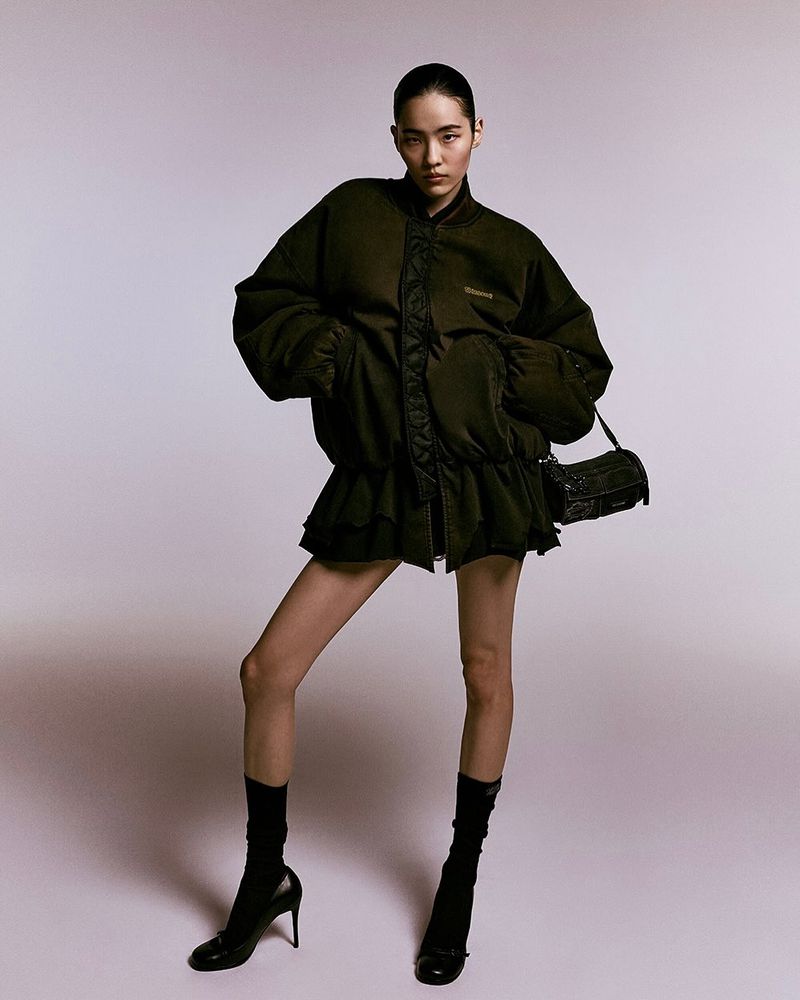 A tall Asian woman poses in a fashion shot, wearing a dark puffer jacket, mini skirt, black heels, and carrying a small black purse.