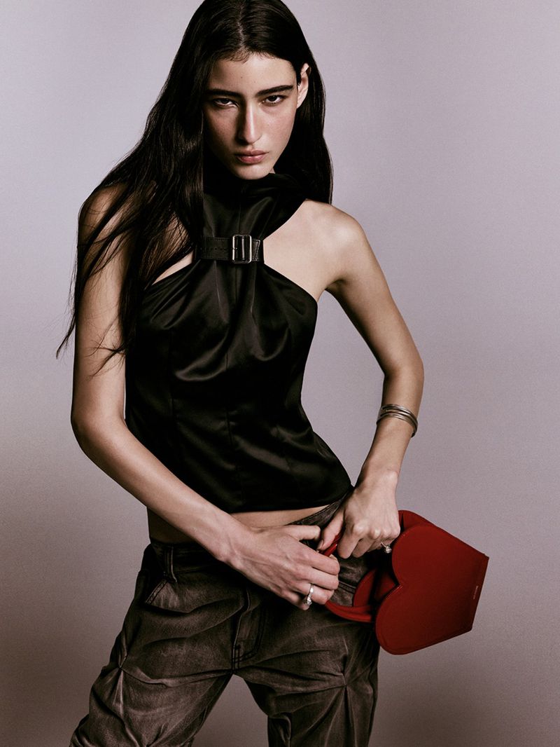 Woman in black halter top and grey pants holds red heart-shaped purse in a studio shot, showcasing fashion and editorial style.