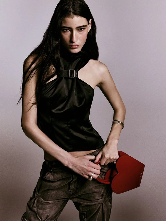 Model in a black halter top and dark trousers holding a red heart-shaped bag against a neutral studio backdrop.