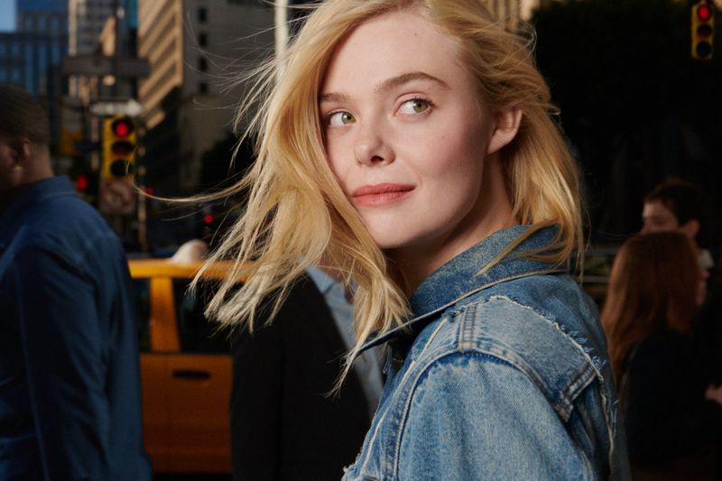 Close-up of a blonde woman in a denim jacket looking off-camera in a city setting with a yellow taxi and pedestrians in the background.