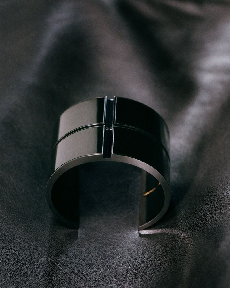 Close-up studio shot of a polished black cuff bracelet with a geometric design, set against a textured black leather background.