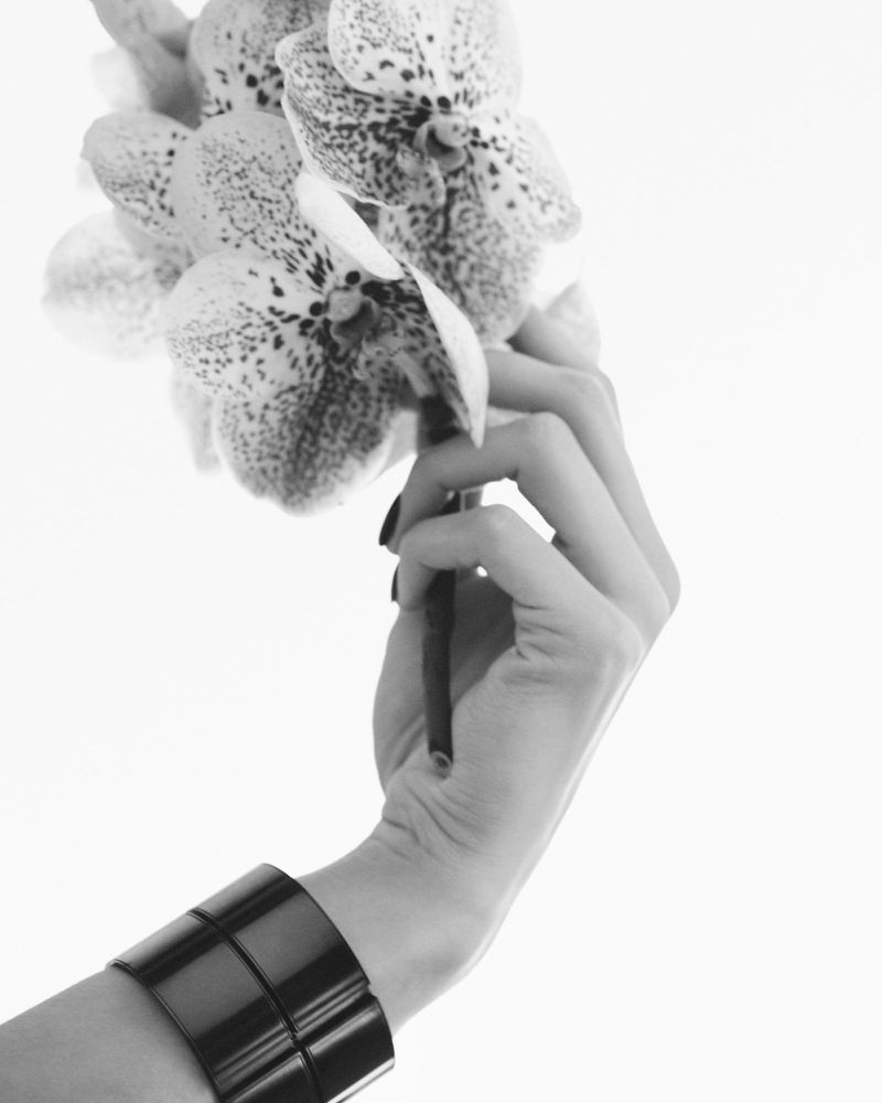 Monochrome image of a woman's hand holding an orchid flower, featuring a black cuff bracelet against a bright white background.