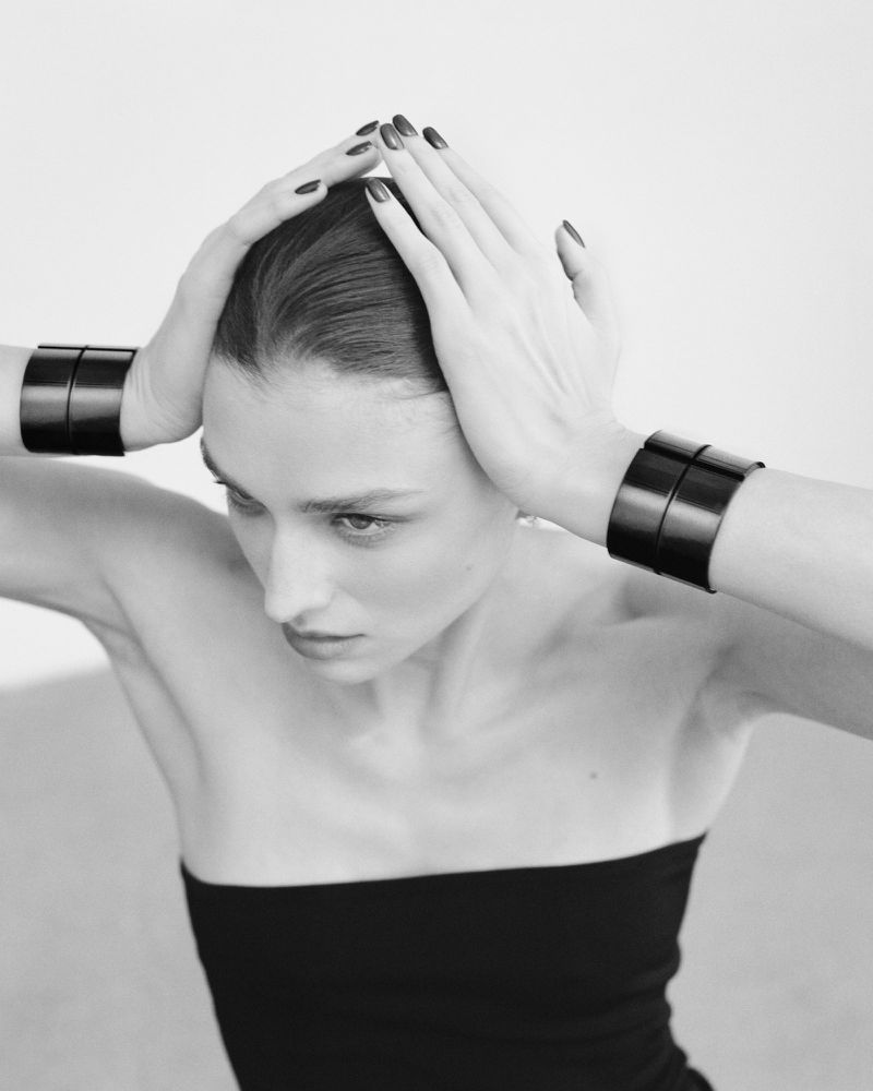 Black and white portrait of a model with black wrist cuffs, hands near her head, and wearing a black strapless top against a plain background.