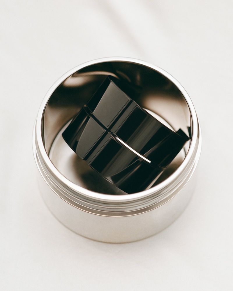 Close-up studio shot of a shiny black square perfume bottle inside a metallic silver container on a white background.
