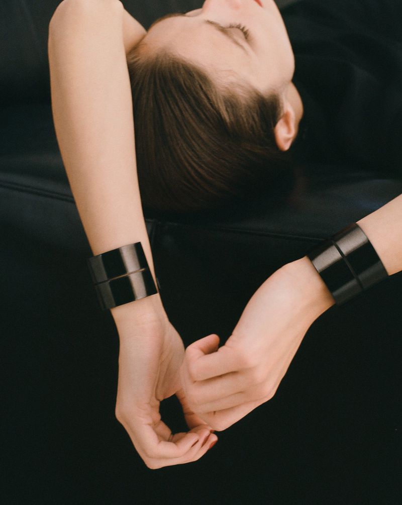 Adult female model lying on a black surface wearing thick dark wrist cuffs in a medium shot with a dark background.