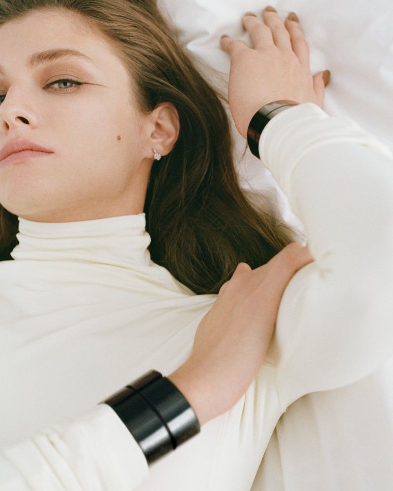 A woman with long brown hair poses against a white background, wearing a white turtleneck sweater and a black cuff bracelet on her wrist.