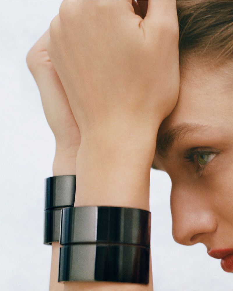 Close-up studio shot of a fair-skinned woman with green eyes and red lipstick wearing shiny black wrist cuffs with a white background.