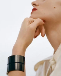 Close-up of a model wearing a wide black cuff bracelet and resting chin on hand against a white backdrop.