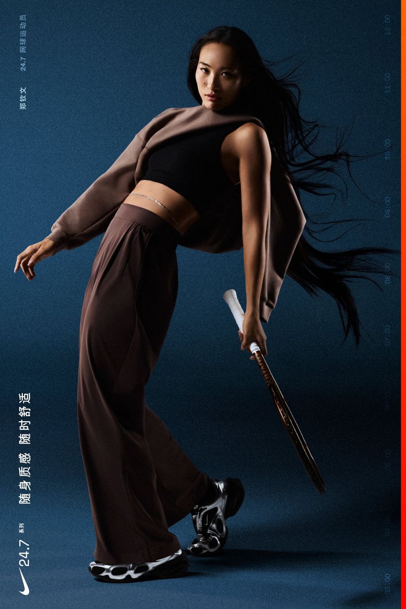 Athletic woman poses in relaxed brown apparel while holding a tennis racket against a dark blue backdrop in a fashion editorial shot.