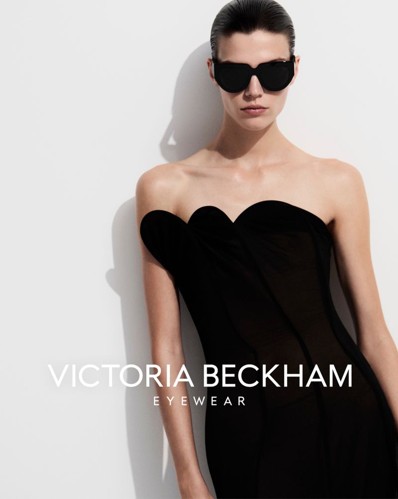 Fashion shot of a model wearing sunglasses and a black strapless dress against a white wall, promoting Victoria Beckham Eyewear.