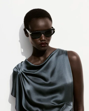 Model wearing dark sunglasses and a slate blue satin top against a clean white seamless studio background.