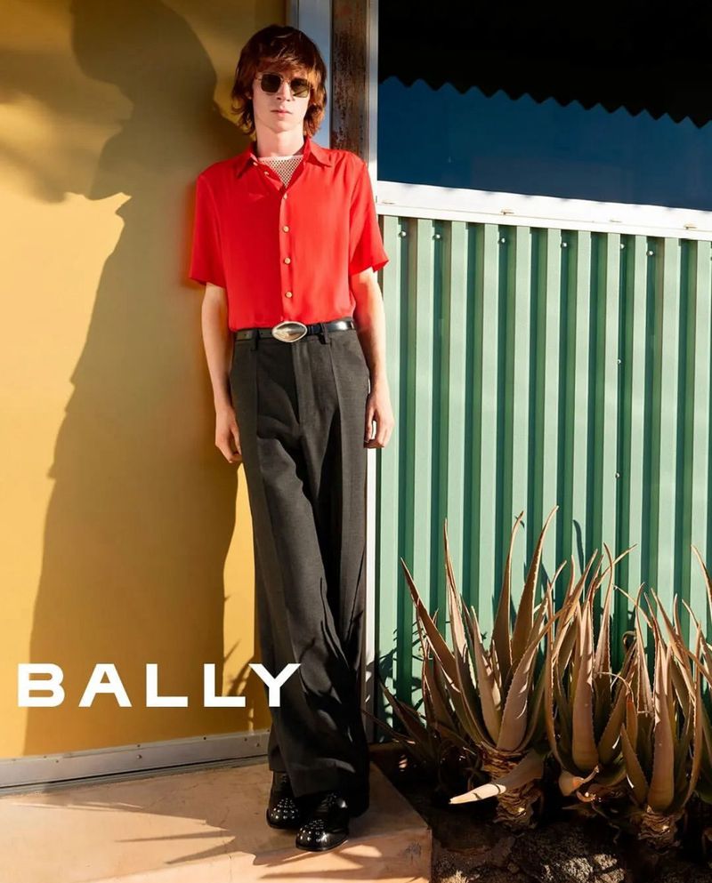 Stylish man in red shirt and black trousers posing against a textured wall with aloe plants, bathed in warm, directional sunlight.
