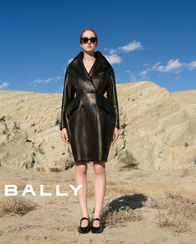 Fashionable woman in a black leather trench coat, posing in a desert with a bright blue sky, and the brand name BALLY displayed.