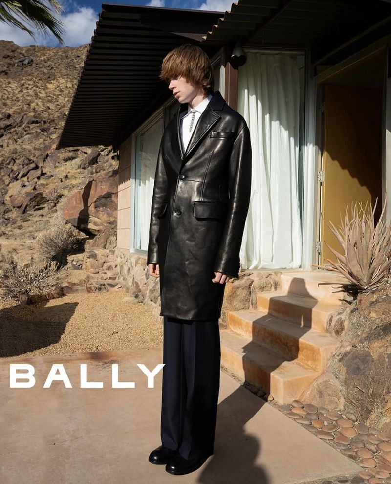 Fashion shot of a male model wearing a black leather trench coat and wide-leg slacks outdoors in front of a modern house and desert landscape.