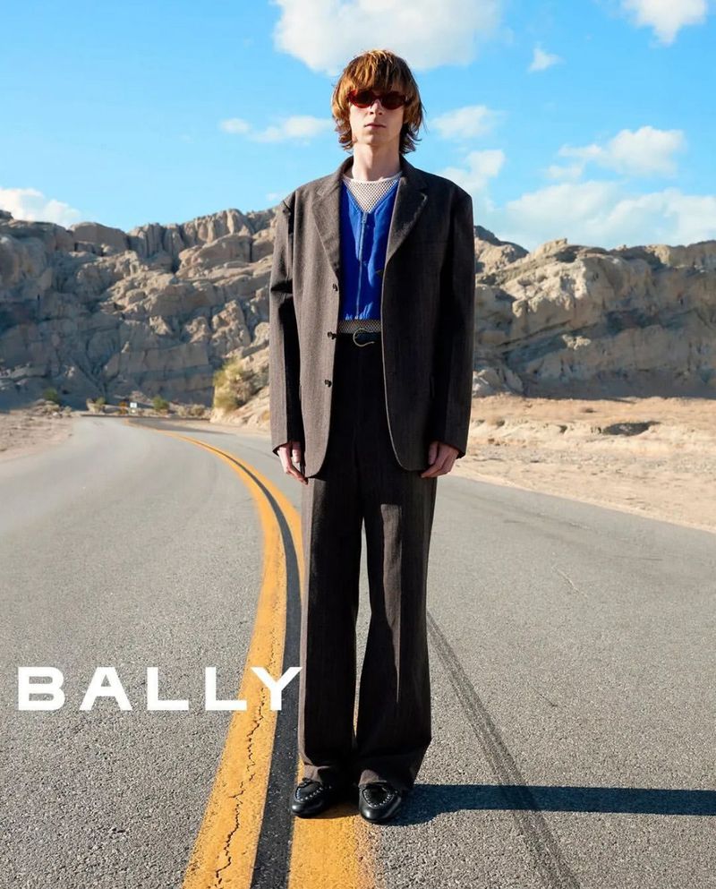 Model wearing a brown suit jacket and trousers standing on an asphalt road with rocky mountain terrain in the background.