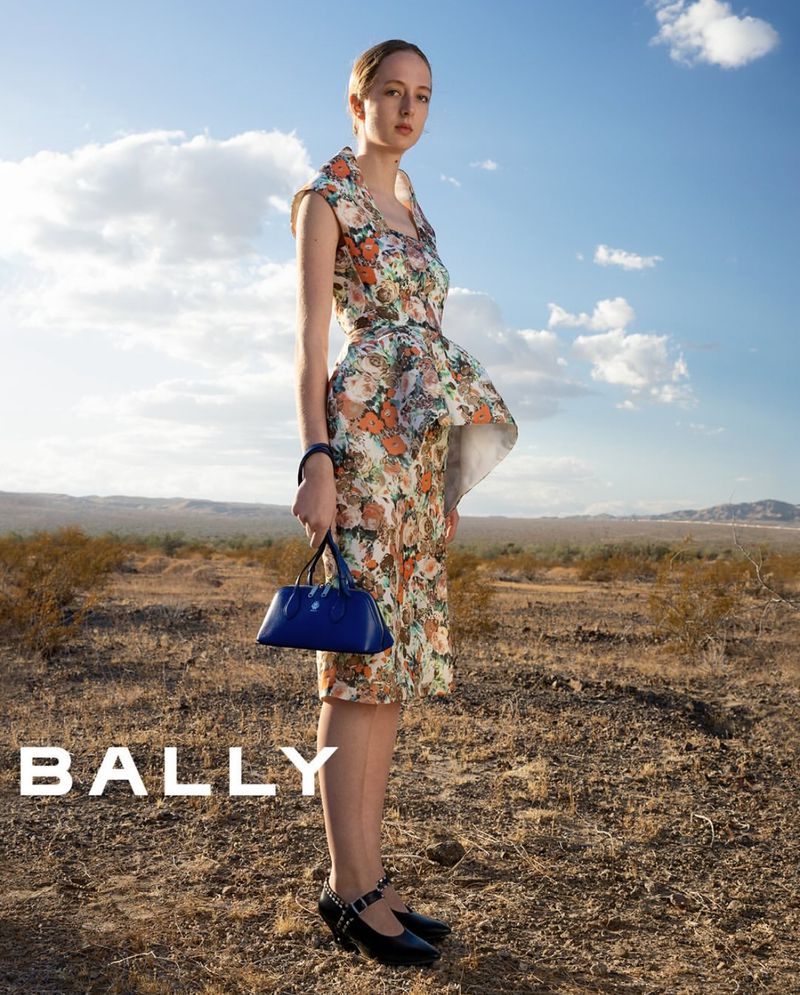 A young woman in a floral dress and black Mary Jane shoes poses with a blue purse outdoors, in a fashion editorial for Bally.