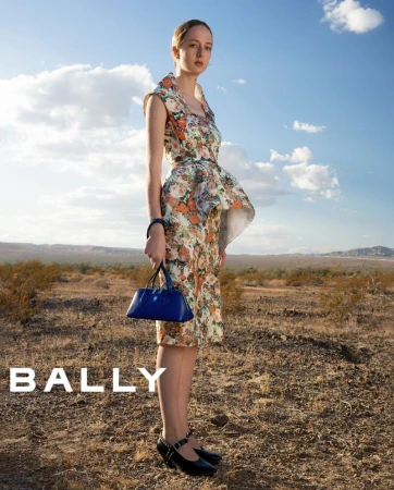 A young woman in a floral dress and black Mary Jane shoes poses with a blue purse outdoors, in a fashion editorial for Bally.