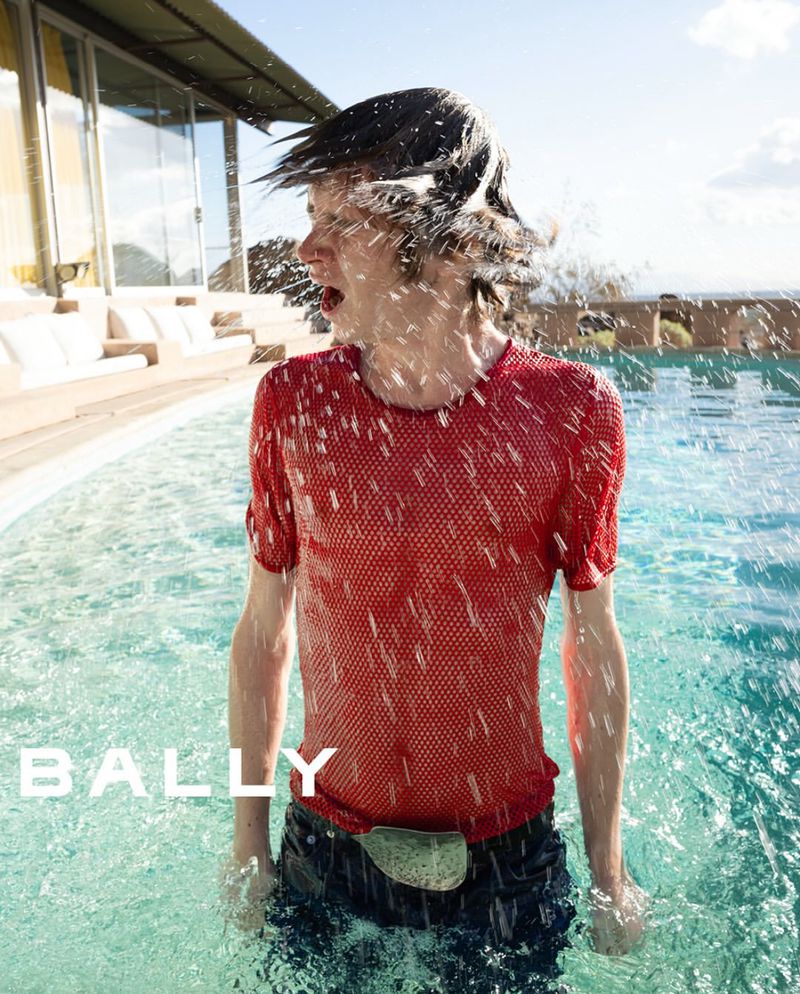 A person in a red shirt is submerged in a turquoise swimming pool, water splashing all around them, with a blurred house in the background.