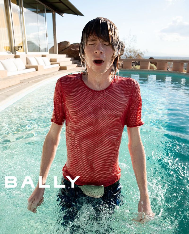 Man in a red mesh shirt emerges from an outdoor pool on a sunny day. Water drips from his face and shirt in a fashion shot.