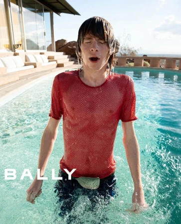 Man in a red mesh shirt emerges from an outdoor pool on a sunny day. Water drips from his face and shirt in a fashion shot.