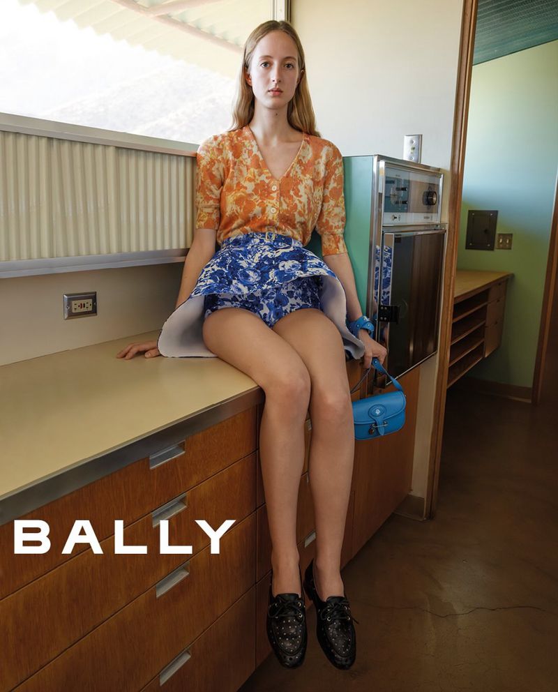 A young woman poses on a kitchen counter in an orange floral sweater and blue skirt, accessorized with a matching blue purse and black loafers.