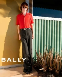 Model wearing a bright red short-sleeve shirt and dark trousers leaning against a yellow wall next to green corrugated paneling.