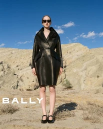 Model wearing dark sunglasses and a structured black leather coat standing in a desert landscape against a blue sky.