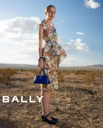 Model wearing a floral print dress and holding a blue handbag in an arid desert landscape under a partly cloudy sky.
