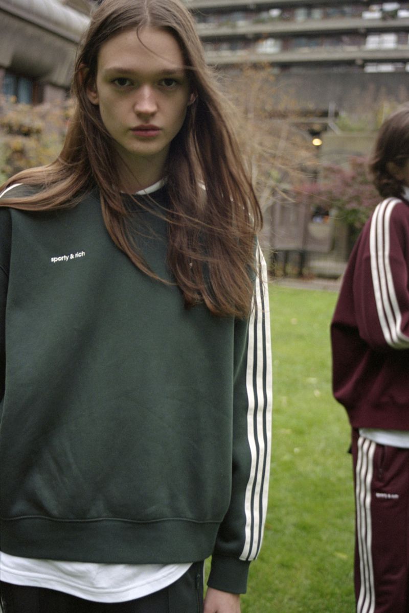Two women in matching tracksuits stand in front of a concrete building in an outdoor setting. The first model is in focus.